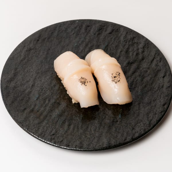 Two pieces of scallop nigiri sushi with black seasoning on a dark textured plate.
