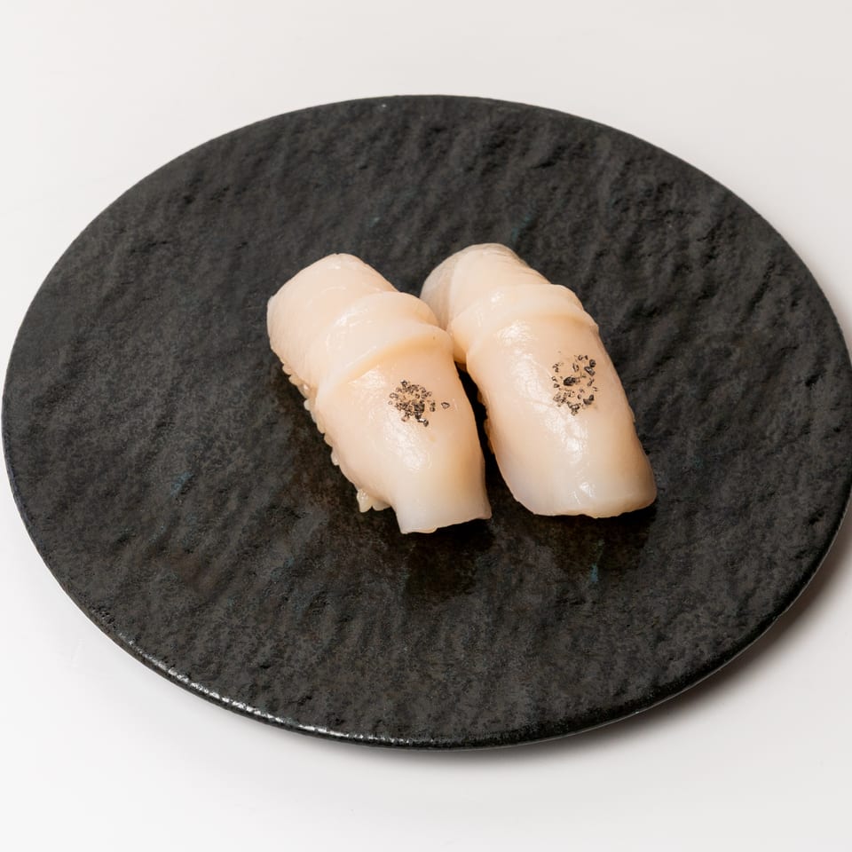 Two pieces of scallop nigiri sushi with black seasoning on a dark textured plate.
