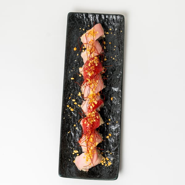 Sushi roll with tuna and salmon, topped with red chili threads and golden flakes on a black plate.