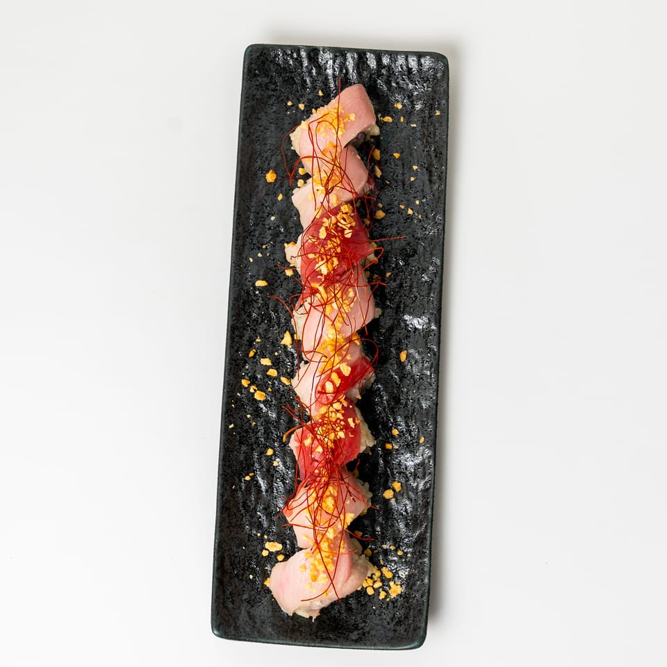 Sushi roll with tuna and salmon, topped with red chili threads and golden flakes on a black plate.