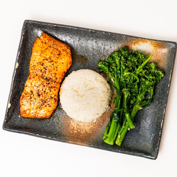 Grilled salmon with sesame seeds, white rice, and broccolini on a rectangular dark plate.