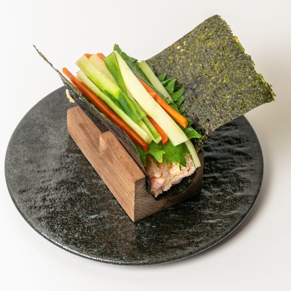A hand roll with crab, rice, and julienned vegetables in a wooden holder on a dark plate.