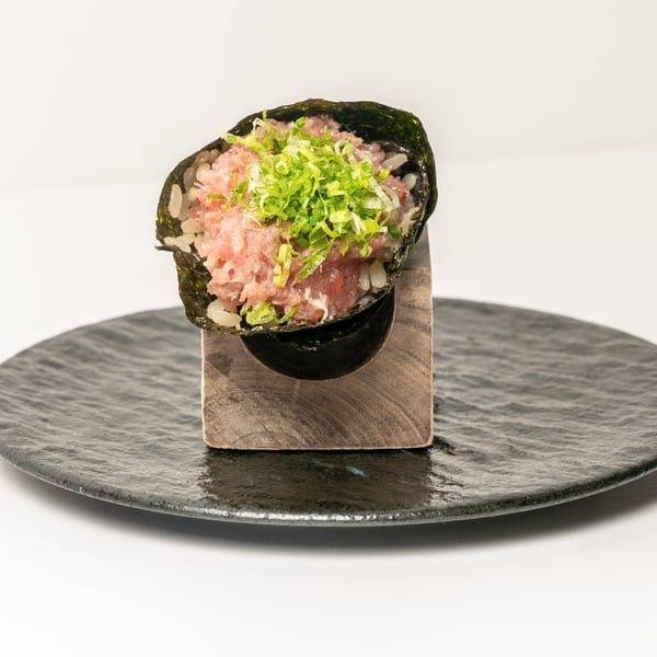 A hand roll with minced tuna, rice, and green onions, served upright on a black plate.