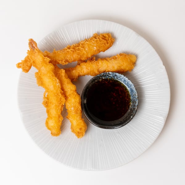 Four golden-brown fried shrimp tempura with a small bowl of dark dipping sauce on a white plate.