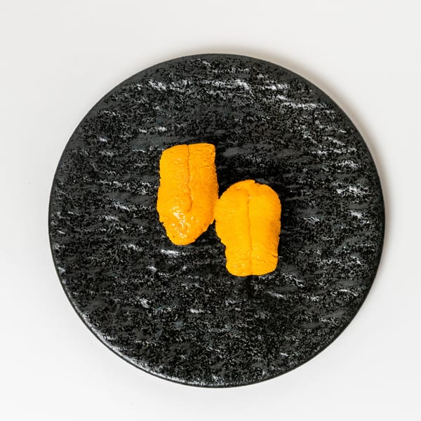 Two bright orange pieces of uni (sea urchin) sit on a textured black oval plate.