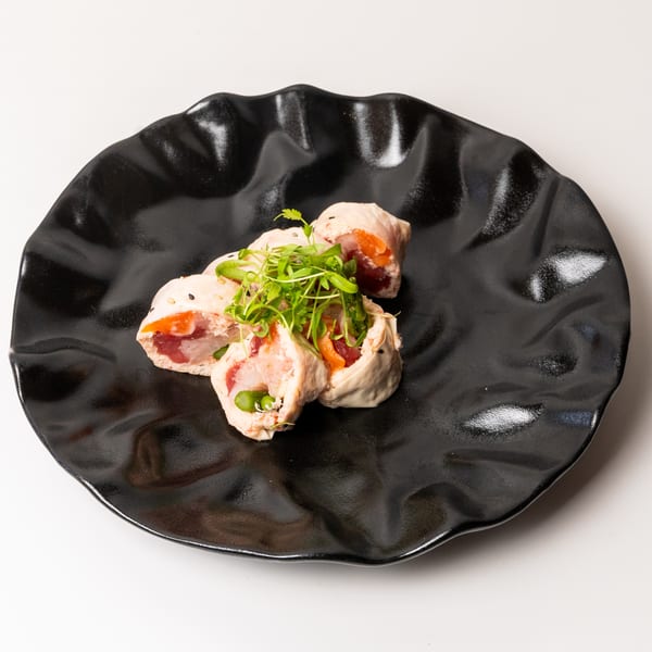Four sushi rolls with fish and vegetables, topped with microgreens, on a black wavy plate.