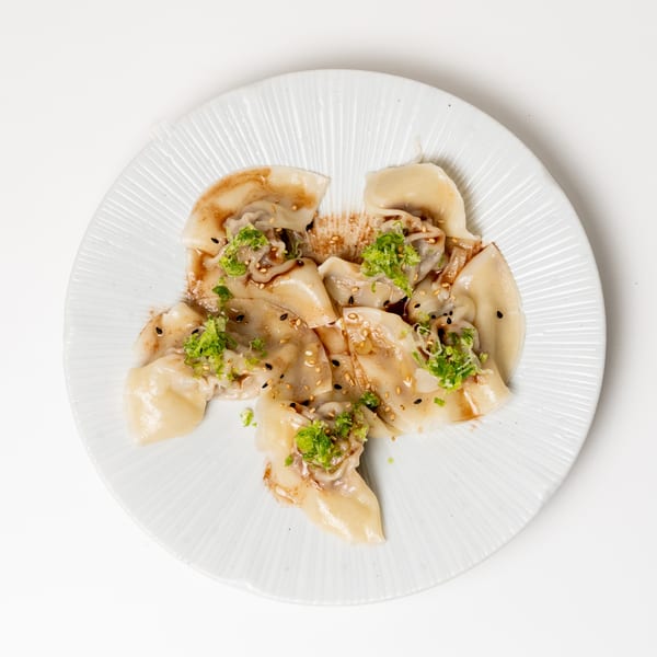 A white plate holds several dumplings drizzled with sauce, topped with green onions and sesame seeds.