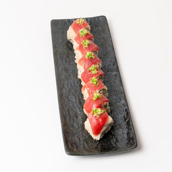 A long, dark rectangular plate holds a row of sushi rolls topped with red tuna and green garnish.