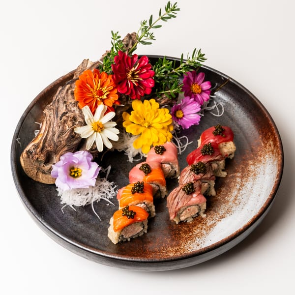 Sushi rolls topped with tuna, salmon, and beef, garnished with caviar, flowers, and wood.