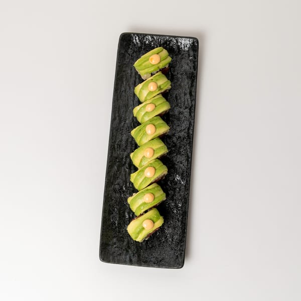 Eight avocado sushi rolls with orange sauce dots, arranged on a black rectangular plate.