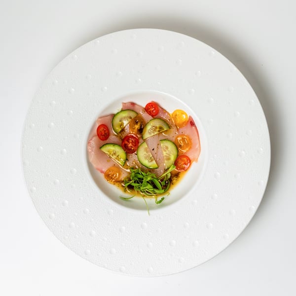 Delicate fish carpaccio with cucumber, cherry tomatoes, and fresh greens on a white textured plate.